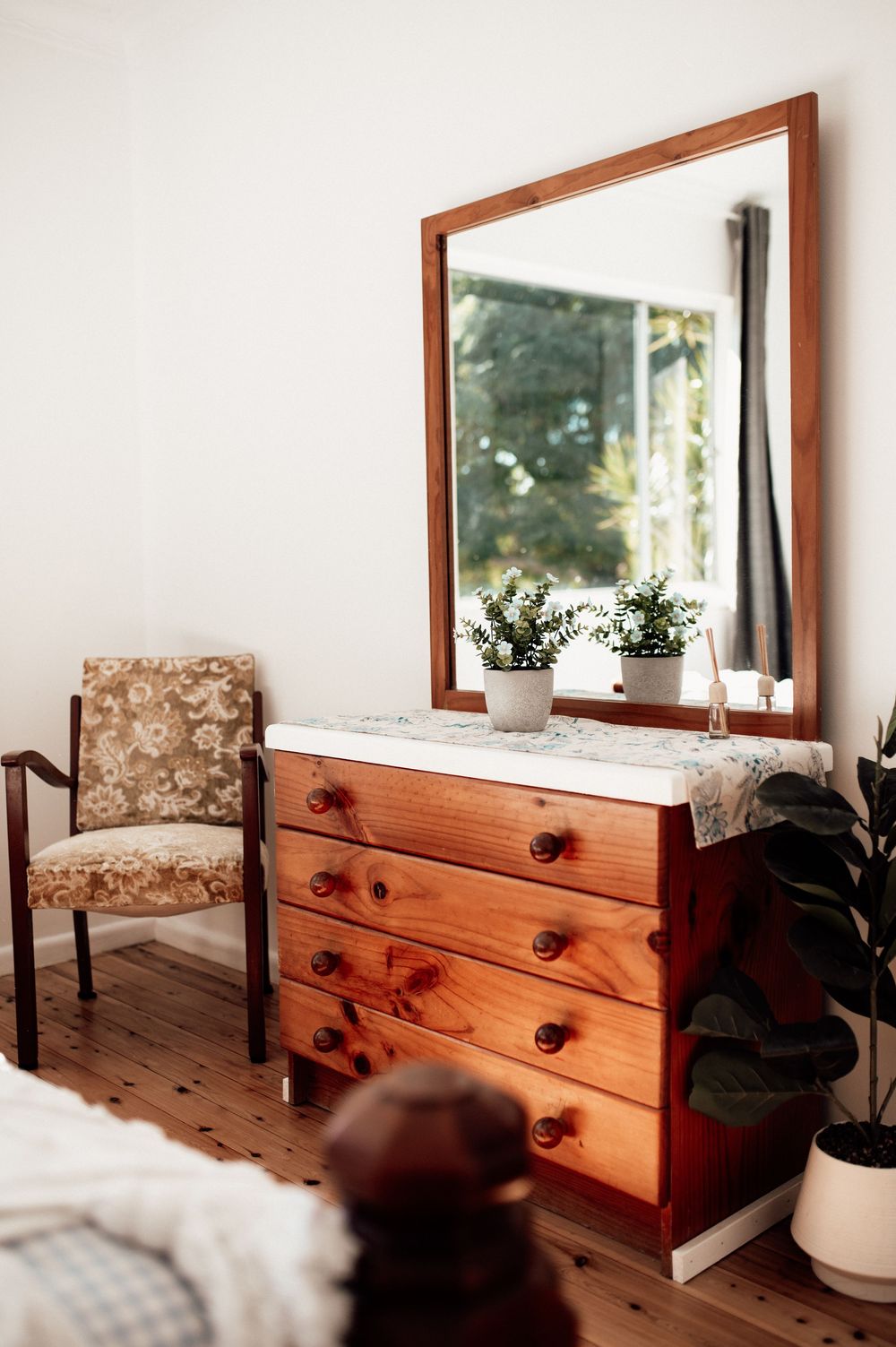 Wooden dresser with mirror and small plants in a cozy bedroom with hardwood floors and a floral armchair.