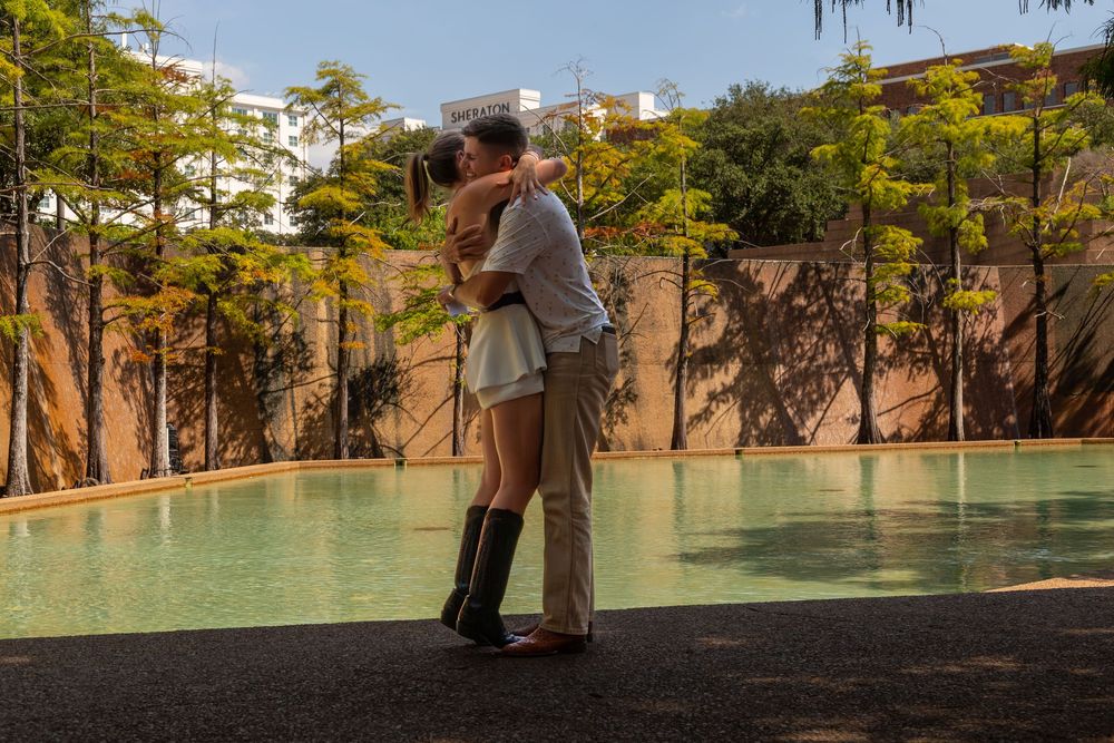 Couple embracing romantically by a serene outdoor pool surrounded by trees and stone walls in warm sunlight.