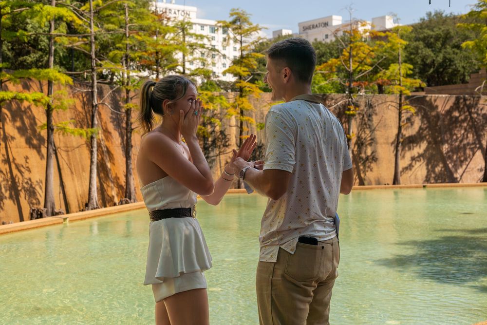 Couple sharing a romantic moment by a serene pool surrounded by lush trees and a rocky backdrop.