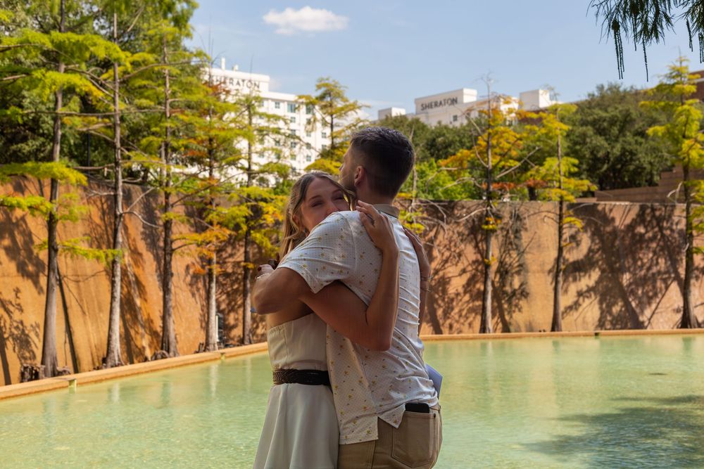 Couple embracing romantically by a serene pool surrounded by lush trees and a modern building in a sunny outdoor setting.