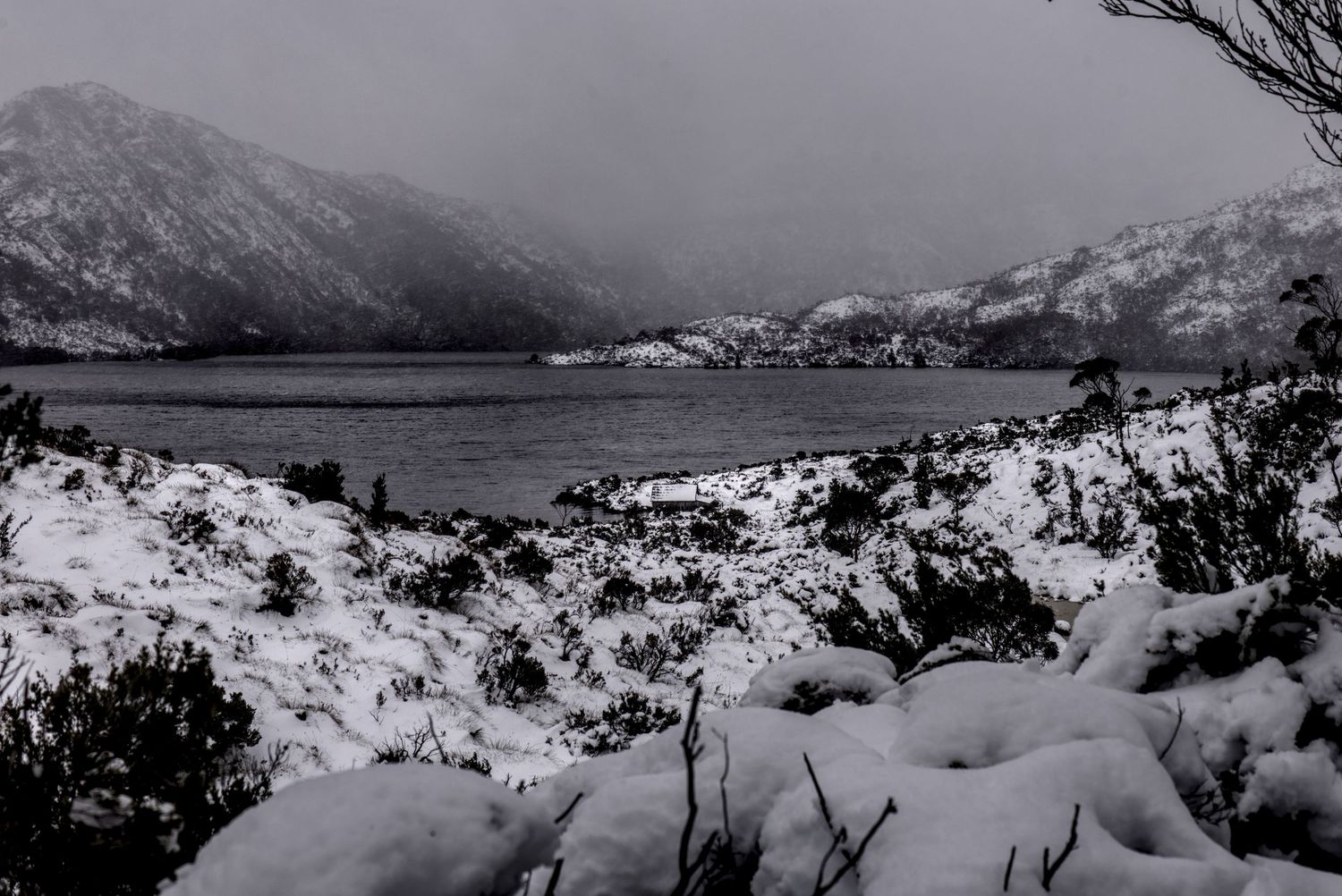 A frozen winter scene of a snow-dusted alpine lake surrounded by dark mountain ranges in monochrome tones.