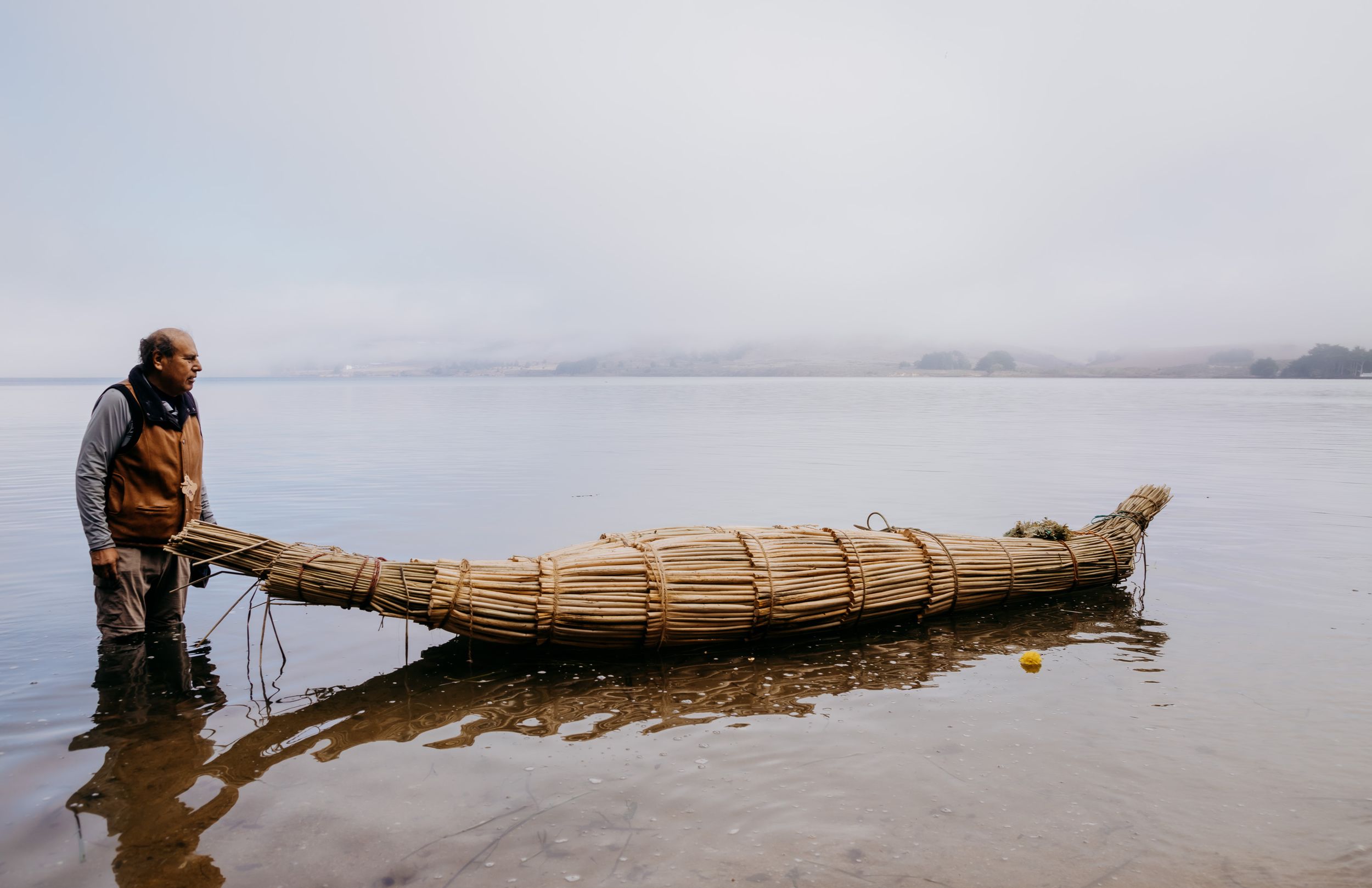 Man standing beside a traditional reed boat on a calm misty lake, showcasing ancient watercraft craftsmanship.