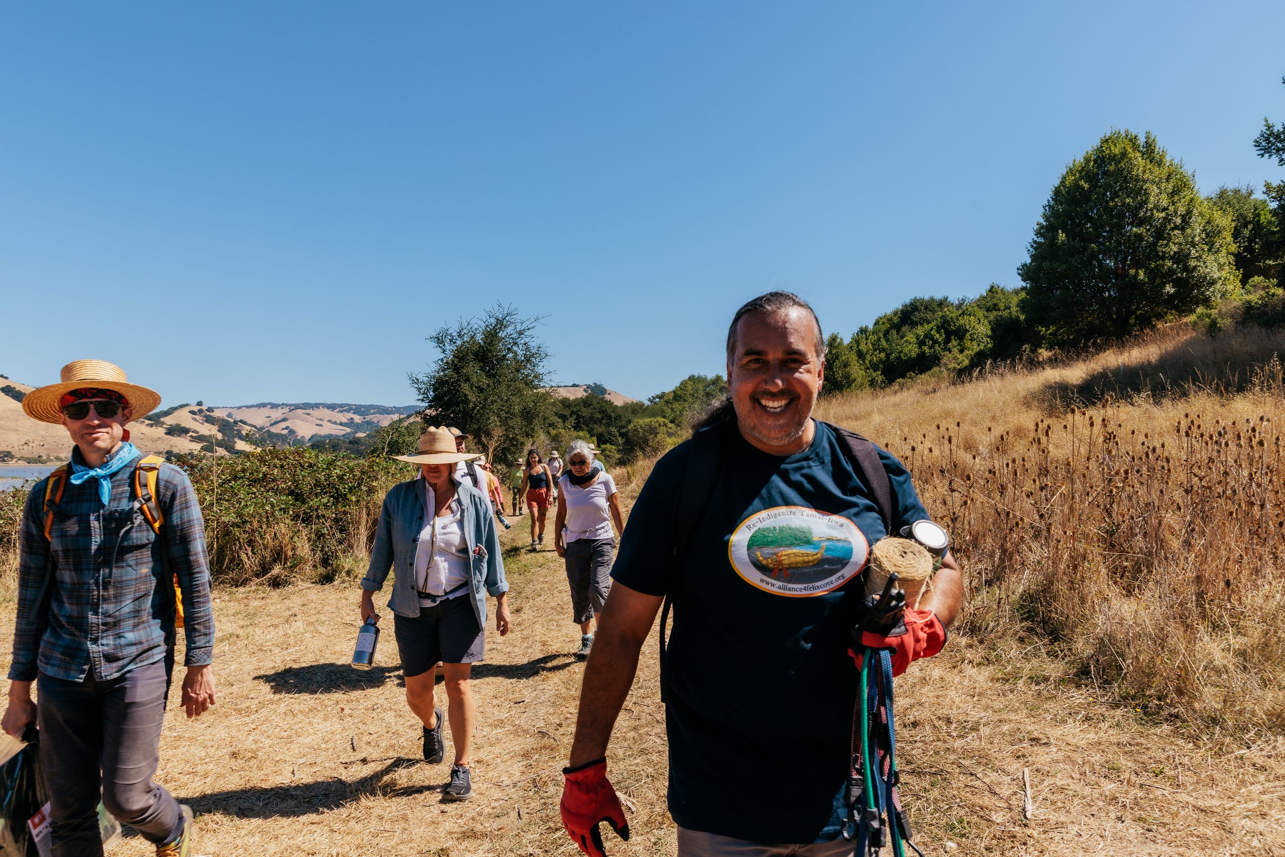 Hikers walking along a sunny trail through dry grasslands with trees and hills in the background.