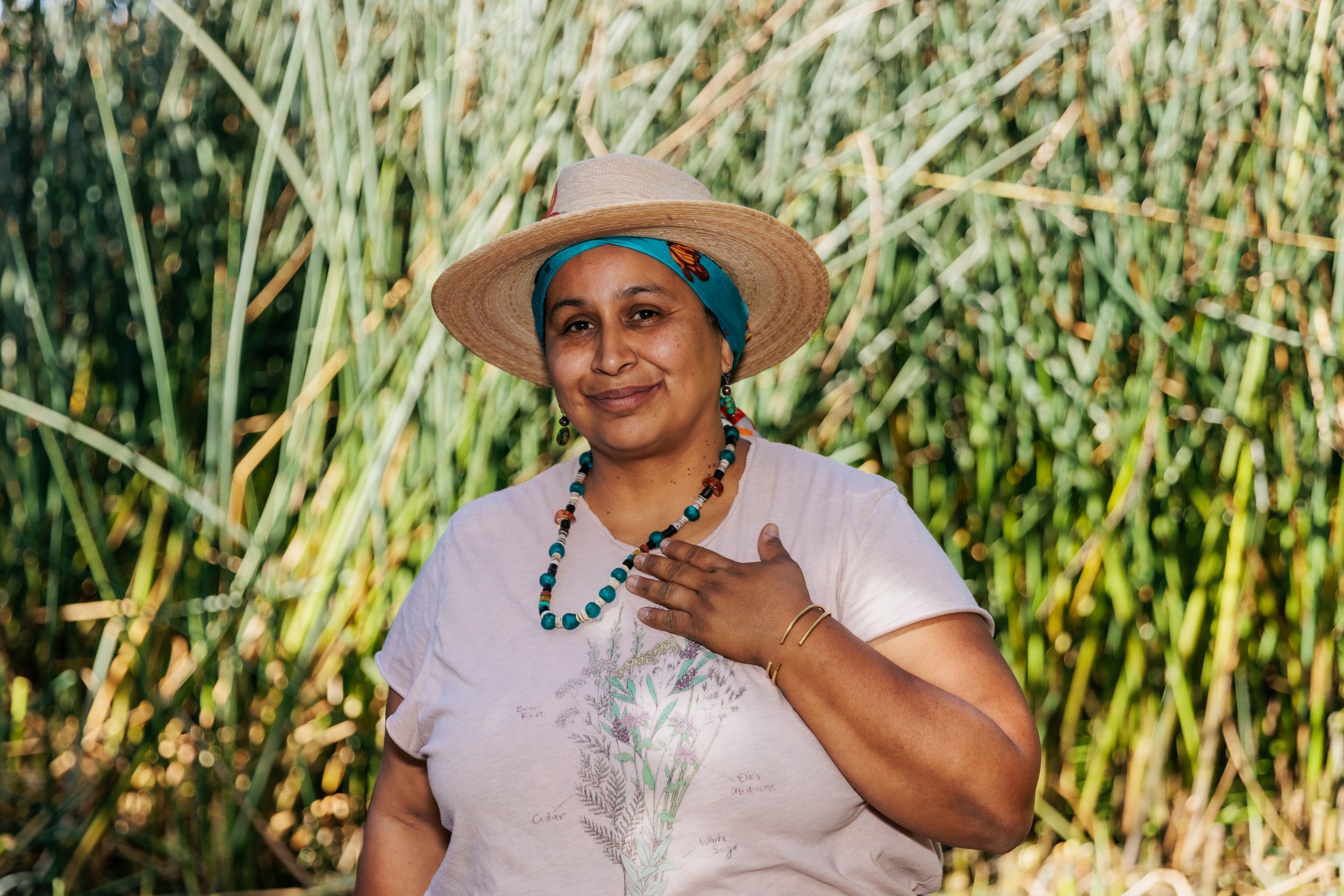 Indigenous woman in straw hat and turquoise necklace smiling outdoors among tall green reeds.