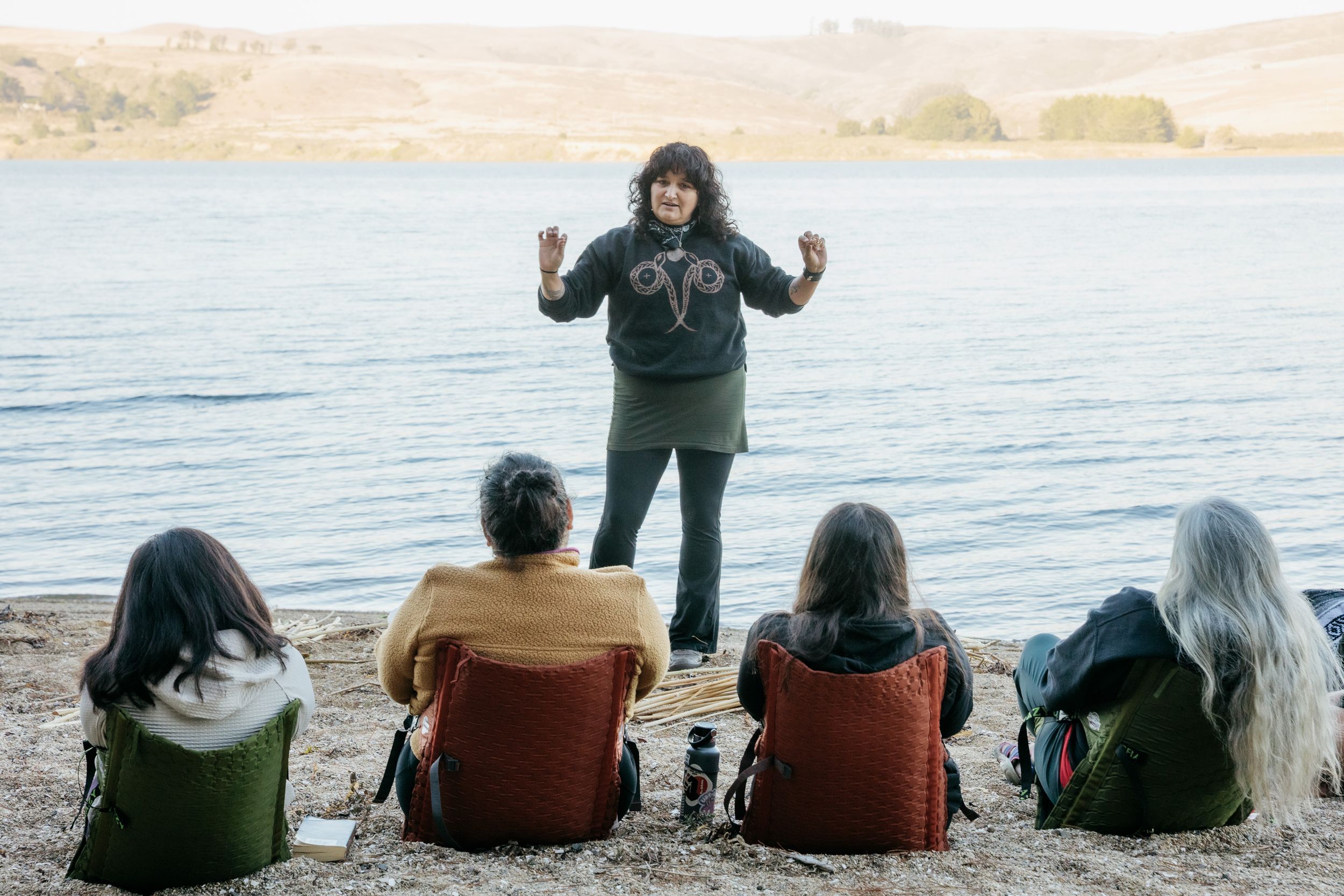 Woman speaking to seated group by lakeside during outdoor educational gathering on sandy shore.