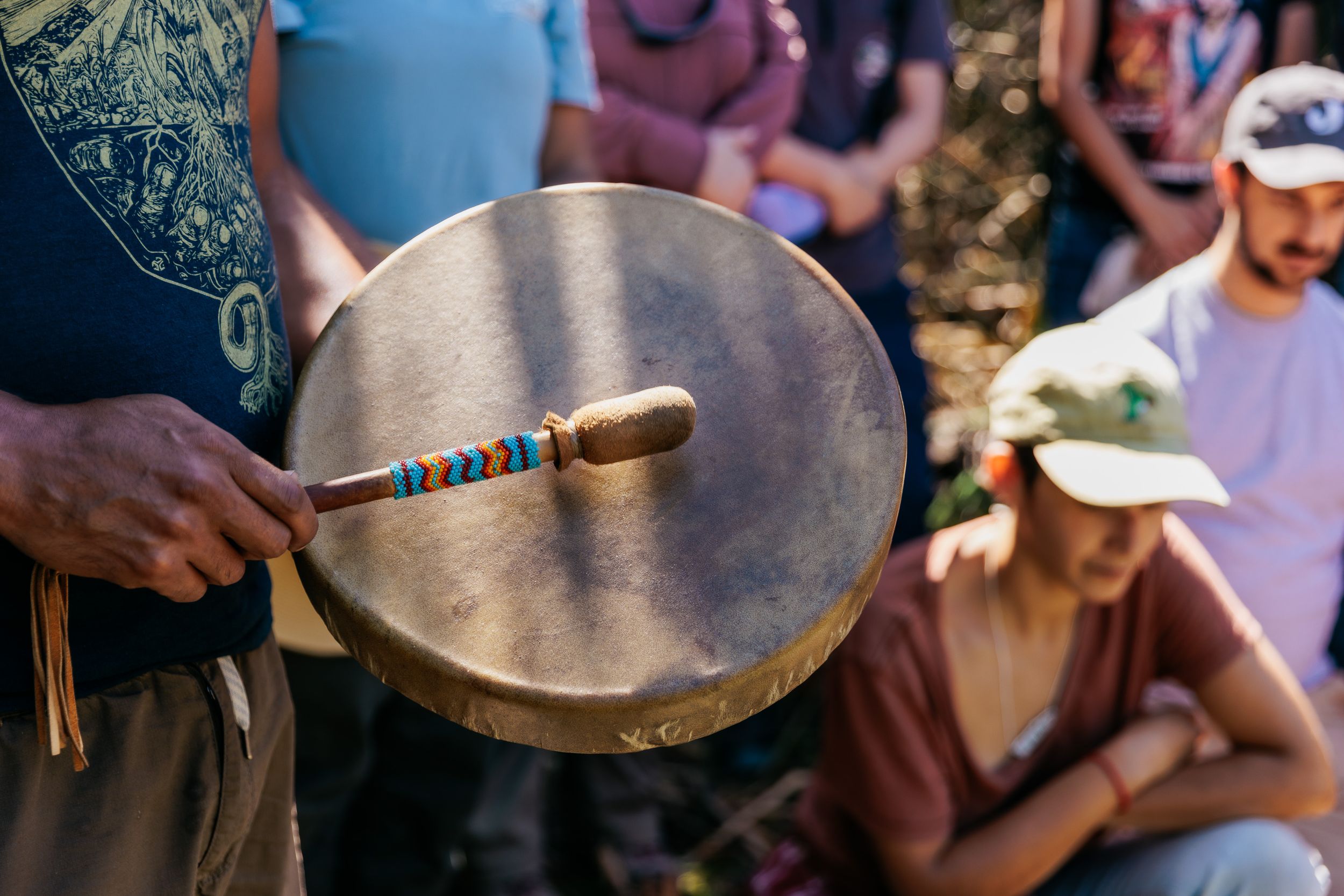 Person playing a traditional Native American hand drum with a colorful beaded drumstick outdoors.