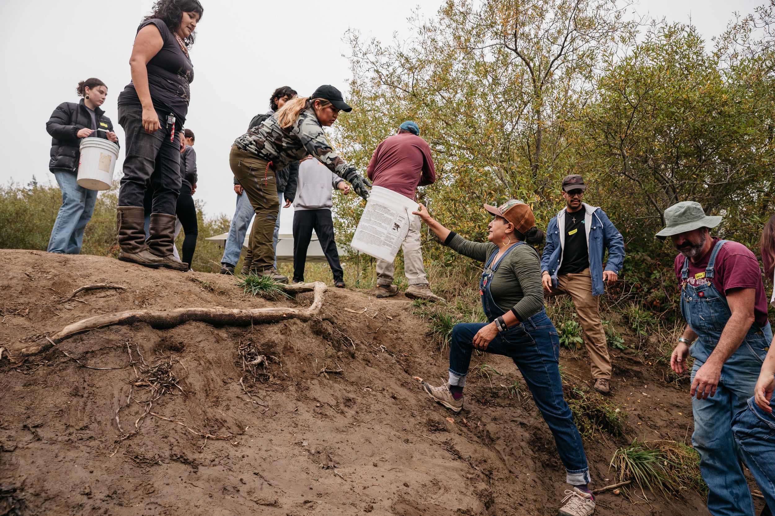 A diverse group of people pass buckets along a muddy hillside during an outdoor restoration project.