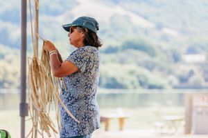 A woman in a blue hat weaving natural fiber rope with a scenic lake and hills in the background.