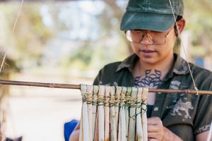 Tattooed craftsperson in green cap carefully arranges bamboo tubes on a wooden frame outdoors.