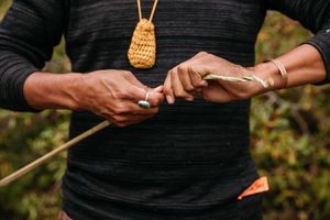 Person in black top crafting a wooden arrow, wearing turquoise ring and gold bracelet with woven pouch necklace.