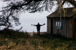 Silhouetted person stands with arms outstretched near rustic wooden shed overlooking misty ocean coastline.