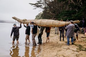A group of people carry a large handcrafted tule reed boat toward the water on a foggy shoreline.