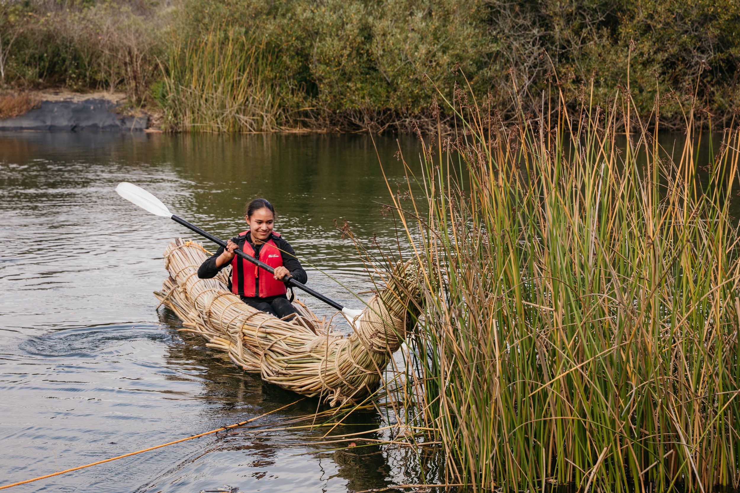 Person paddling a traditional woven reed boat through a marshy waterway wearing a red life vest.
