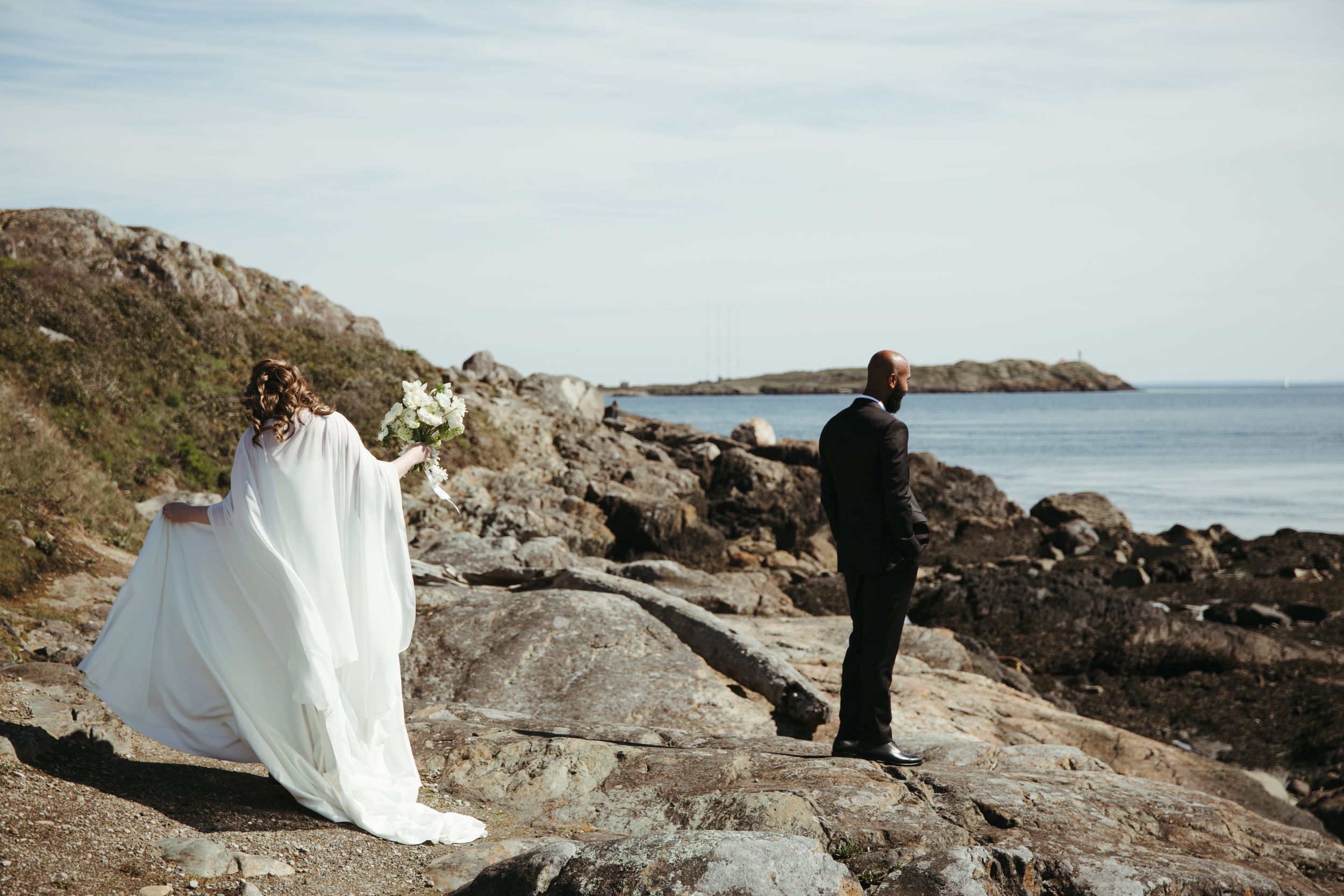 Bride in flowing white cape gown approaching groom on rocky coastal cliffs overlooking calm ocean.