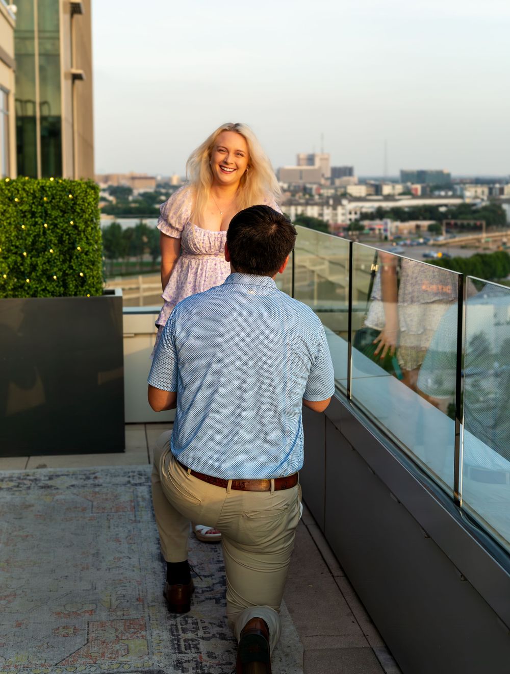 Man kneeling in marriage proposal to blonde woman on urban rooftop terrace at golden hour.