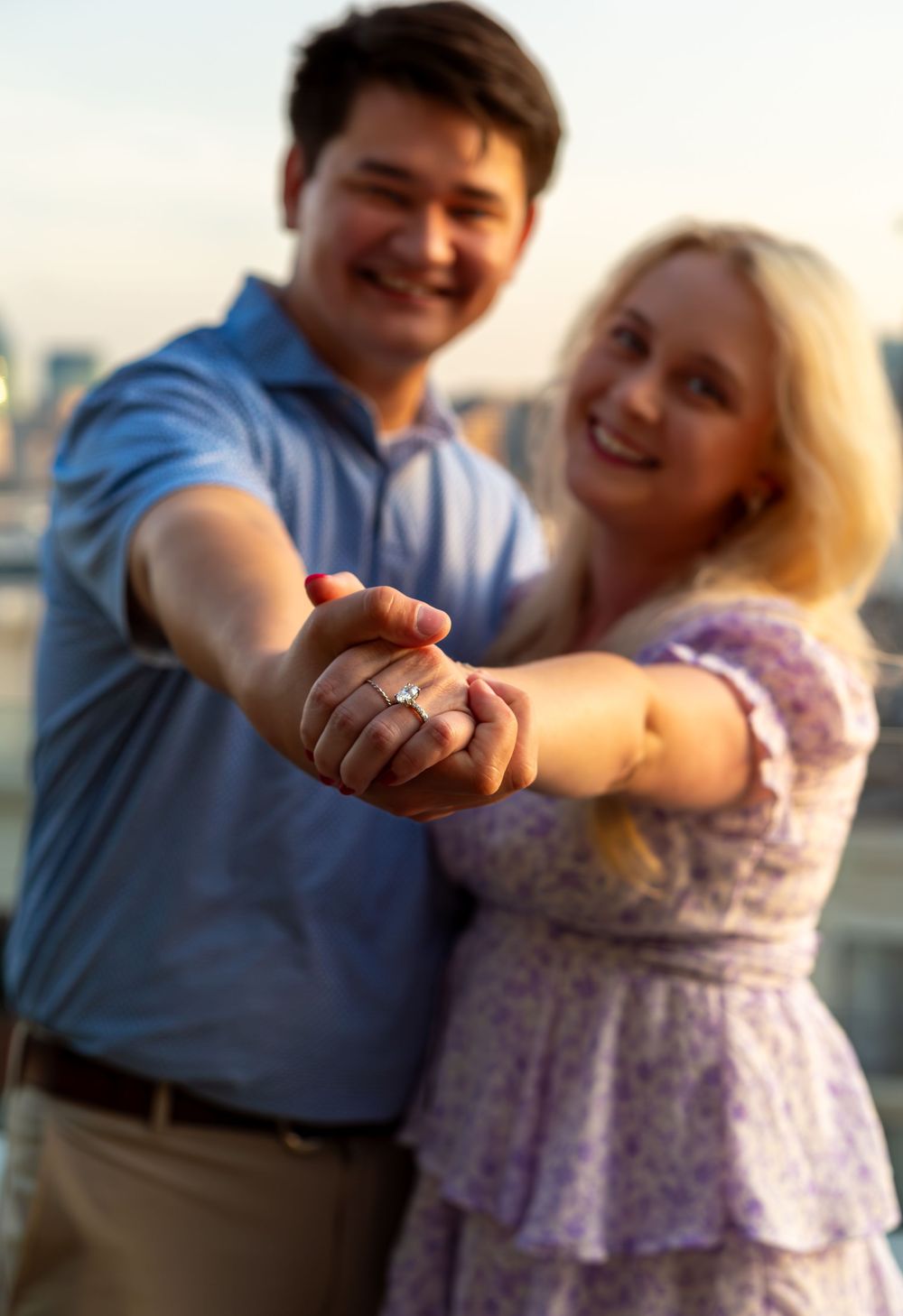 Smiling couple showing off engagement ring outdoors at golden hour with city skyline in background.