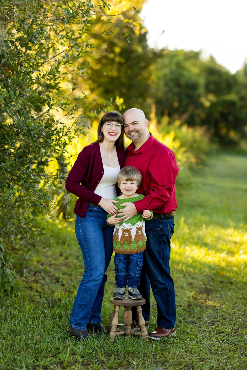 Family of three posing outdoors in red outfits, parents smiling with young child on wooden stool surrounded by green trees.