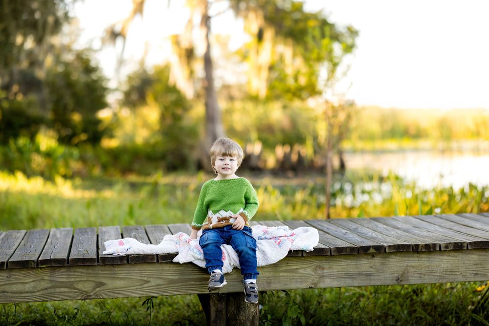 Young boy in green shirt sitting on wooden dock surrounded by lush greenery and Spanish moss-draped trees.