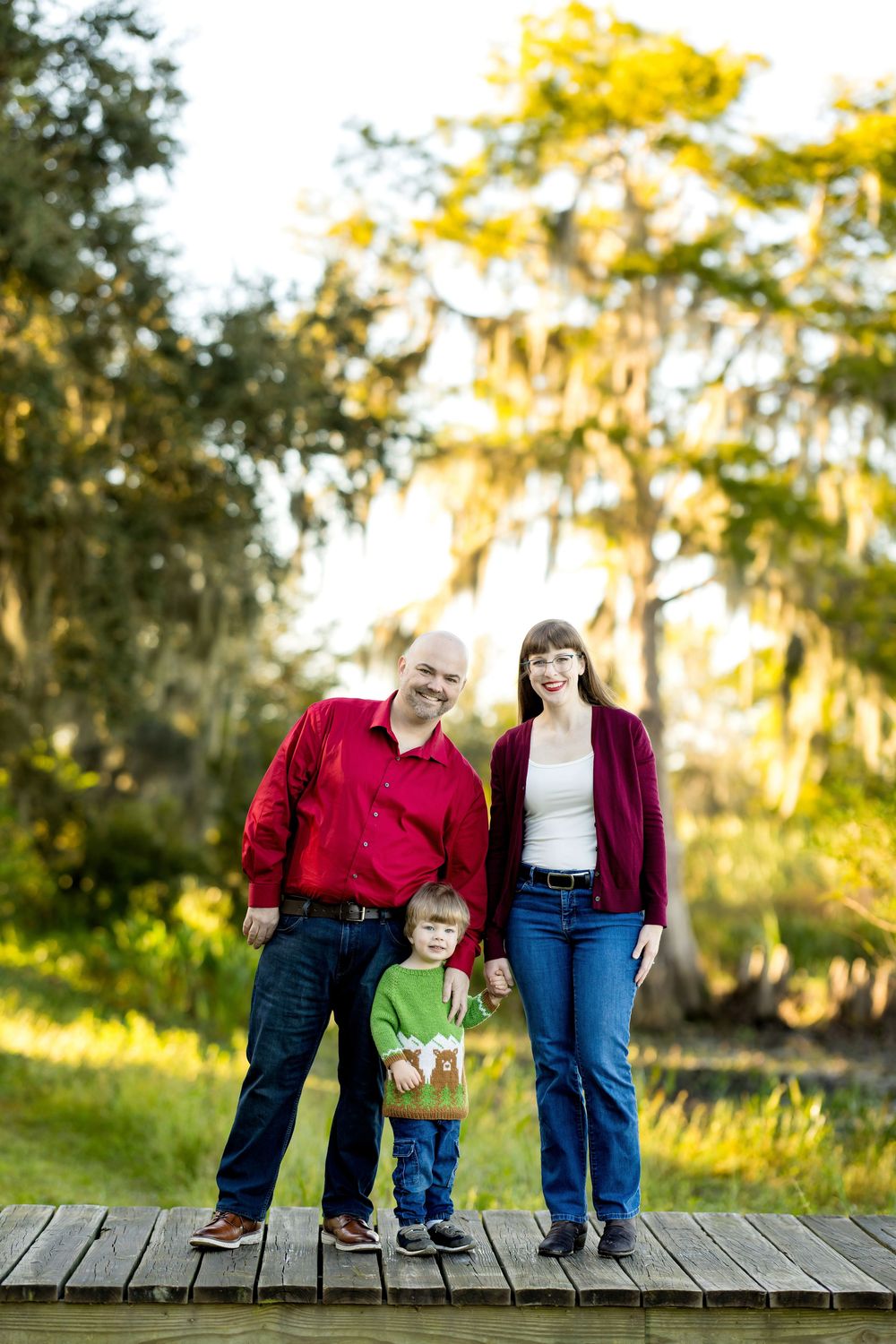 Family of three posing on wooden dock surrounded by lush green trees with Spanish moss.