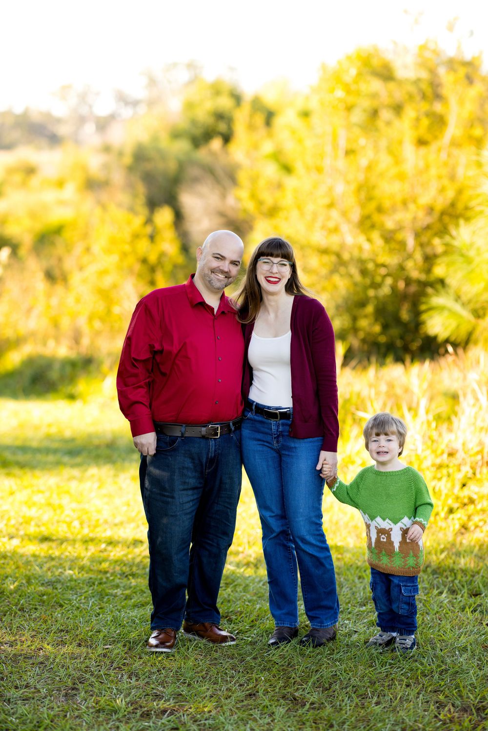 Family of three posing outdoors in fall, parents in red and burgundy with young child in green reindeer sweater.