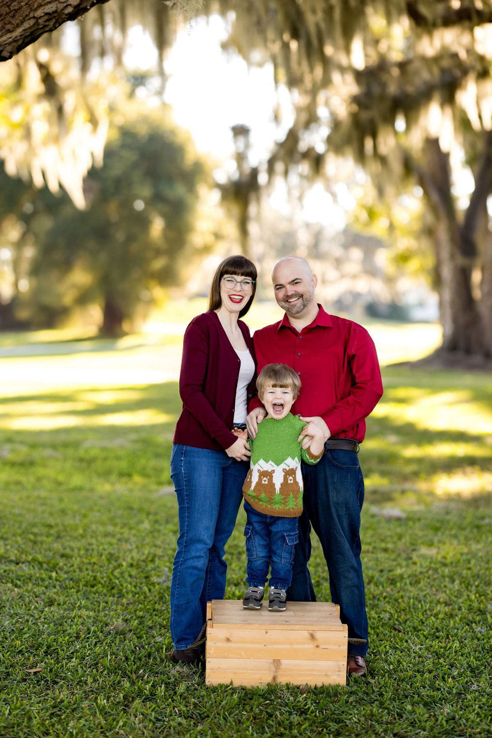 A joyful family of three poses outdoors under Spanish moss-draped trees, wearing festive red and green holiday attire.