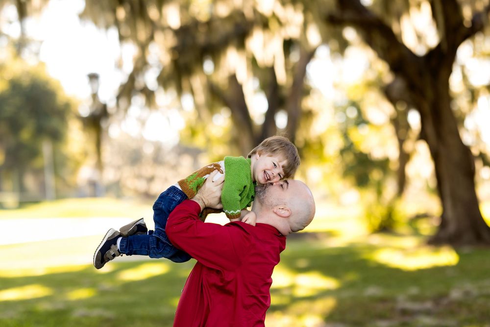 A father in a red jacket joyfully lifts his smiling child in a green outfit outdoors among moss-draped trees.