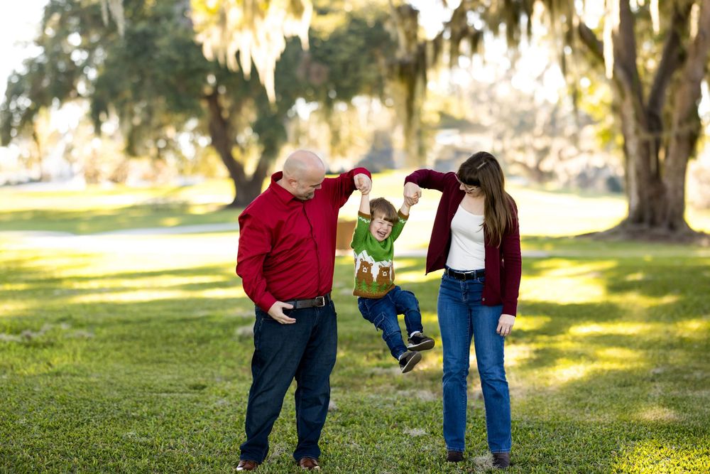 Family of three playing outdoors under moss-draped oak trees, parents swinging young child between them on green lawn.