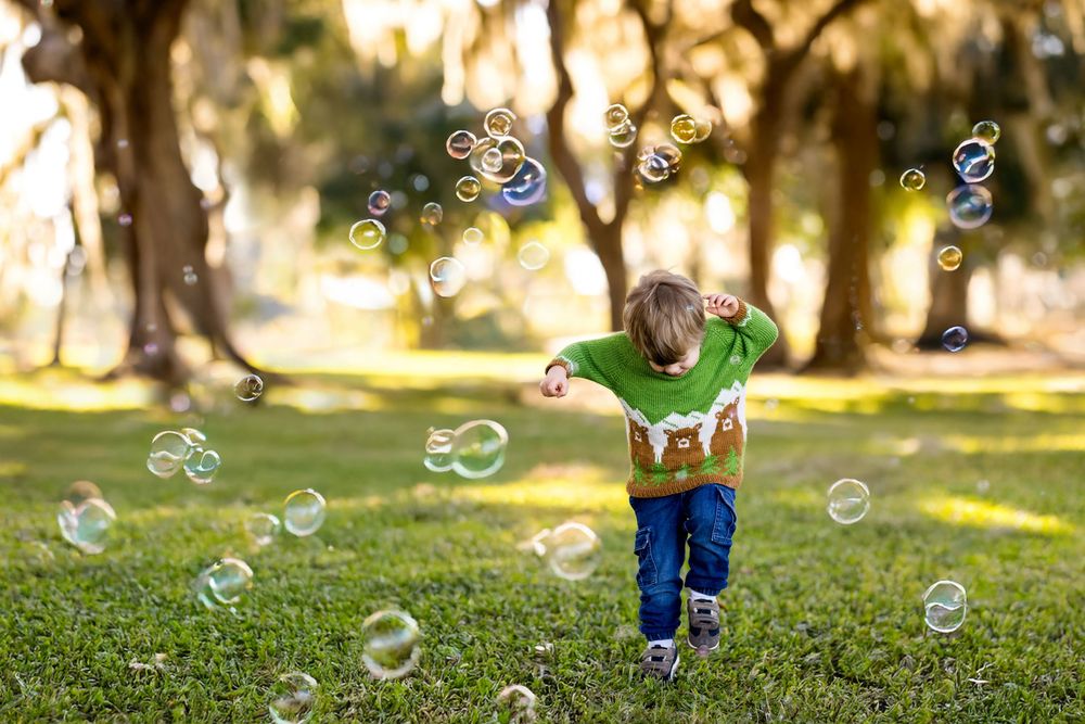 Young boy running through a park chasing colorful soap bubbles on a sunny day.