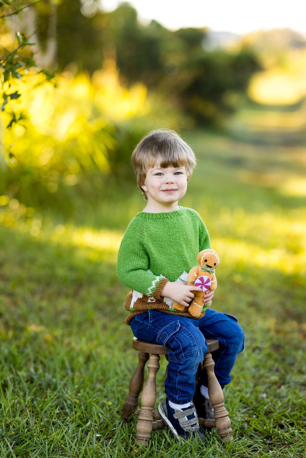 Smiling toddler boy in green sweater and jeans sits on wooden stool outdoors holding a gingerbread man toy.