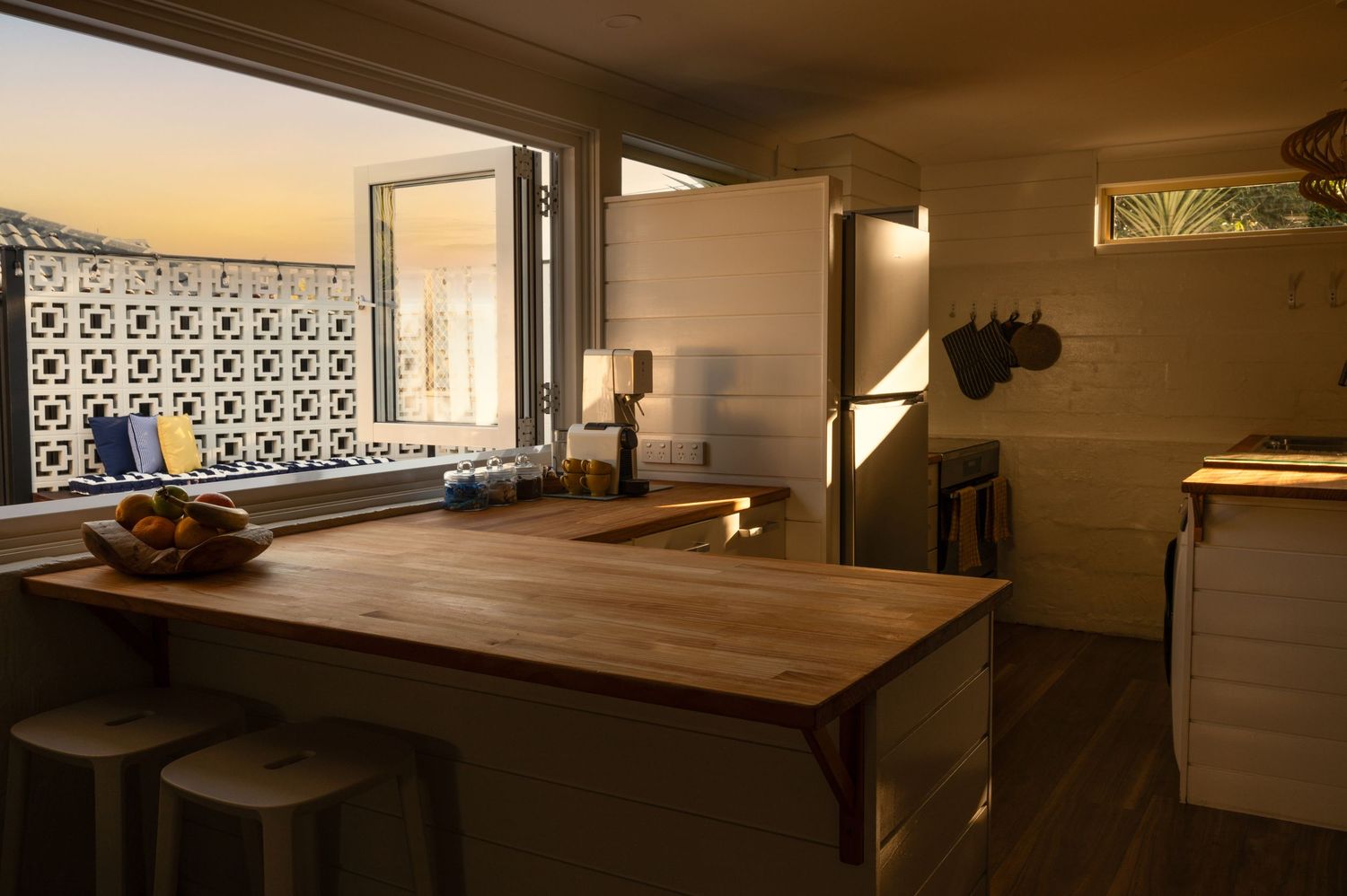 Warm kitchen interior with wooden island countertop, bar stools, and decorative window at sunset.