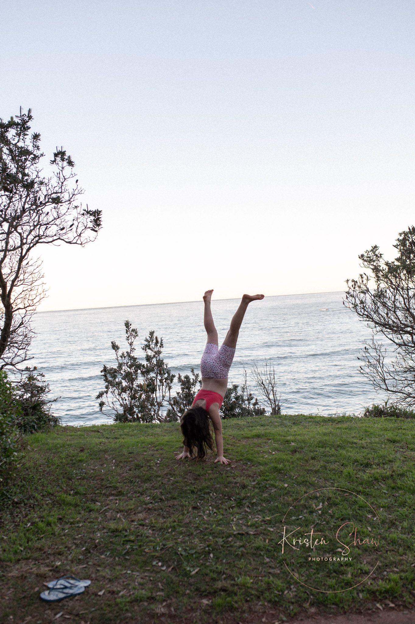 A girl performs a handstand on a grassy hill overlooking a calm ocean at dusk, with trees framing the scene.