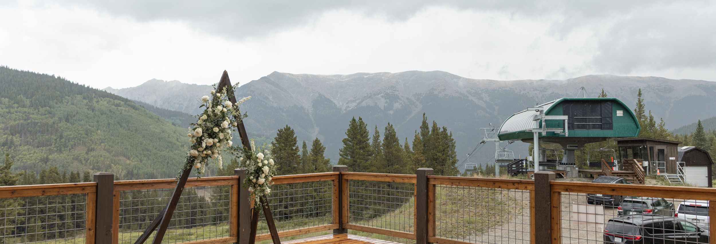 Mountain wedding venue deck with floral arch, panoramic Rocky Mountain views, and rustic wood-and-wire railing under cloudy skies.