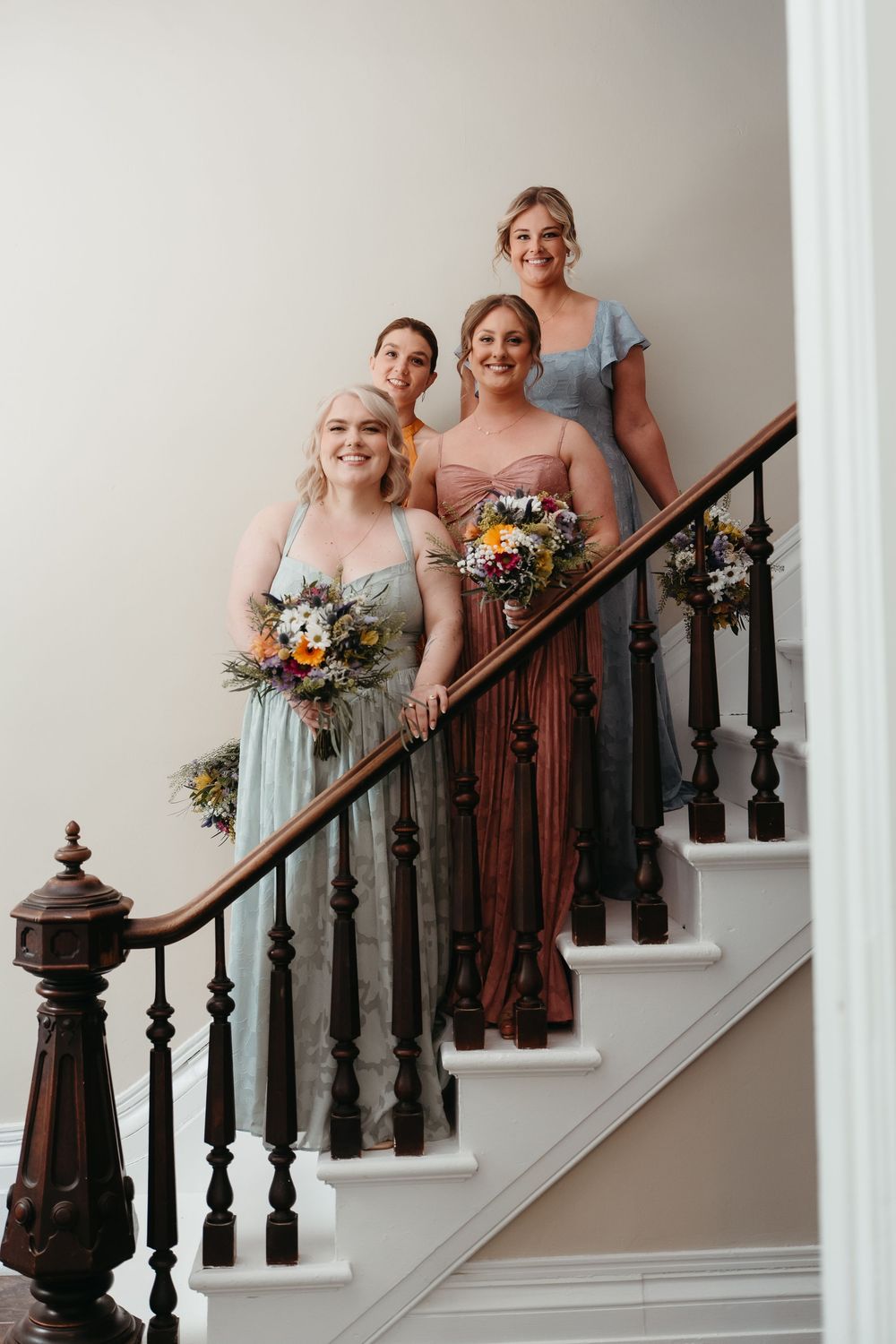 Four bridesmaids in mismatched pastel gowns holding wildflower bouquets pose on an elegant wooden staircase.