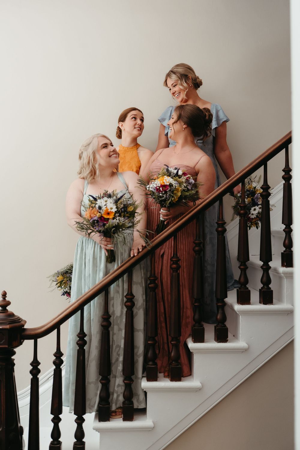 Four bridesmaids in mismatched colorful gowns holding floral bouquets posed on an elegant staircase.