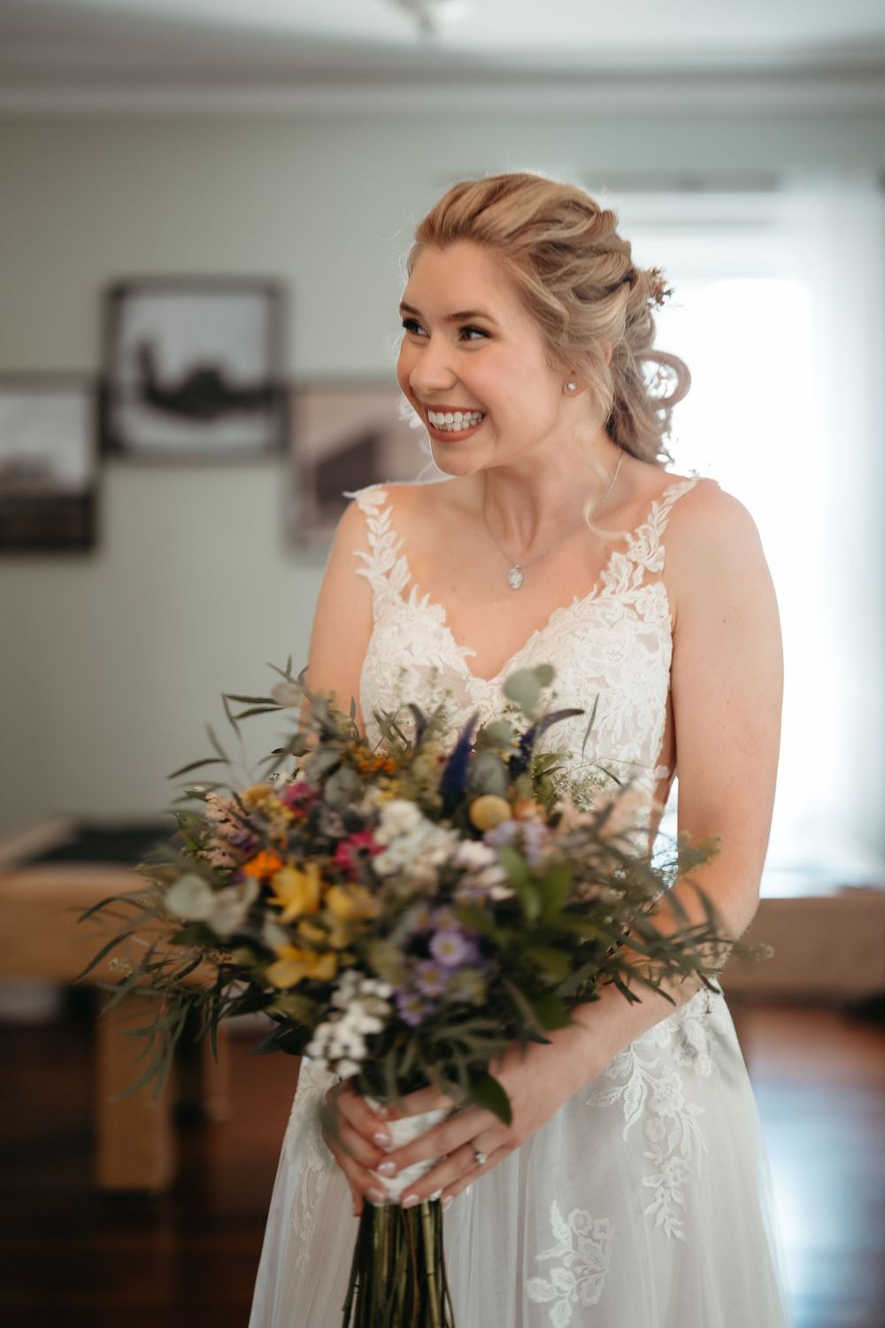 Smiling bride in lace wedding gown holding a colorful wildflower bouquet indoors on her wedding day.
