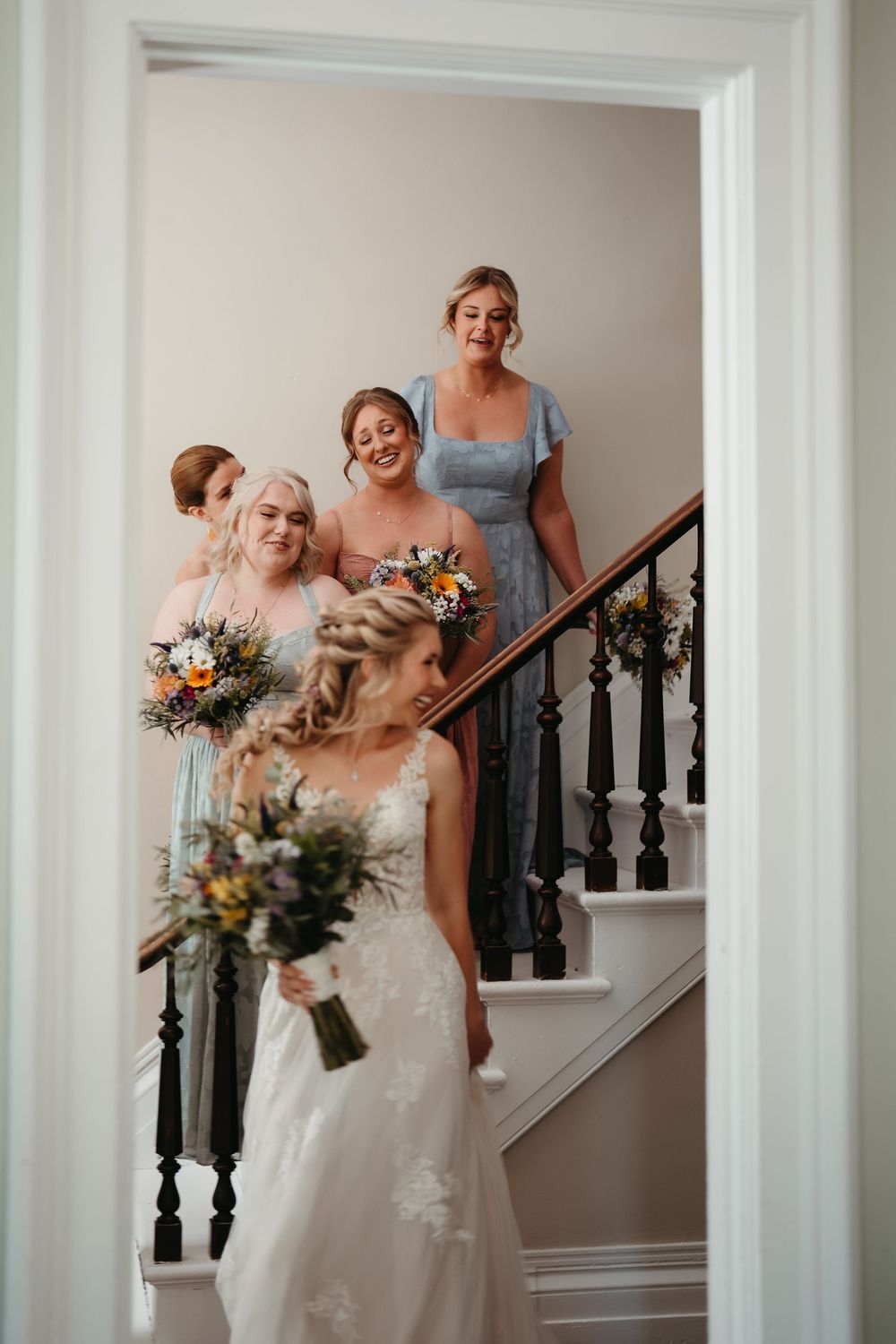 Bride in white lace gown stands at staircase bottom as bridesmaids in colorful dresses descend holding bouquets.