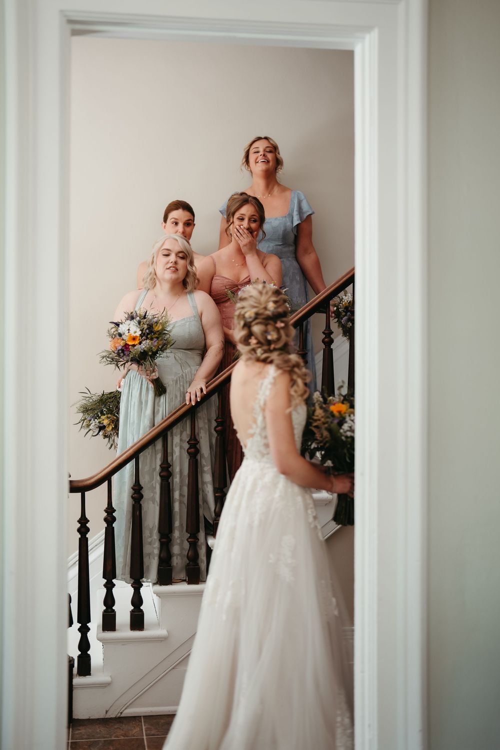 Bride in lace gown reveals look to emotional bridesmaids in pastel dresses on a staircase holding colorful bouquets.