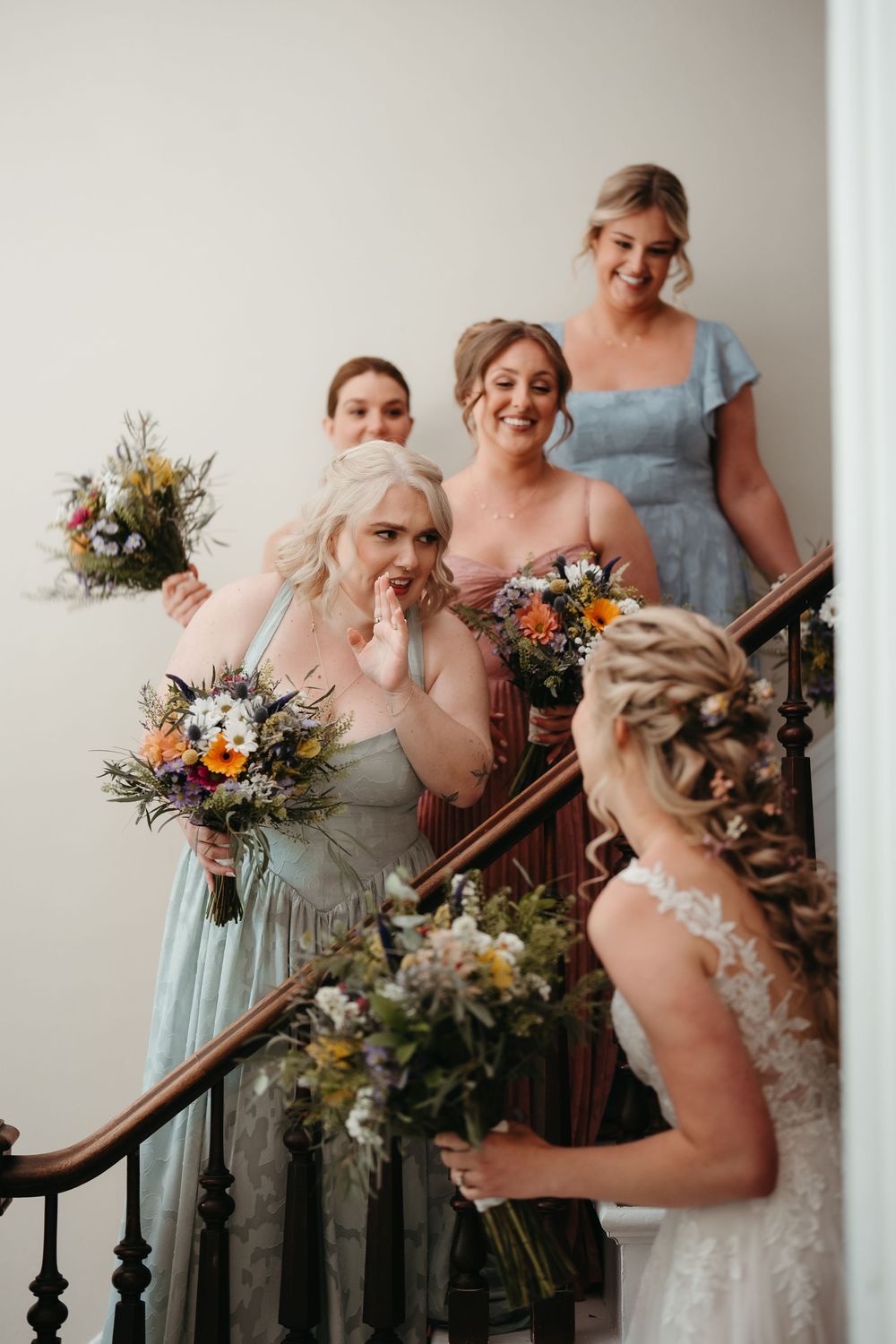 Bridesmaids in blue and pink dresses holding wildflower bouquets react emotionally as bride reveals her wedding gown on staircase.