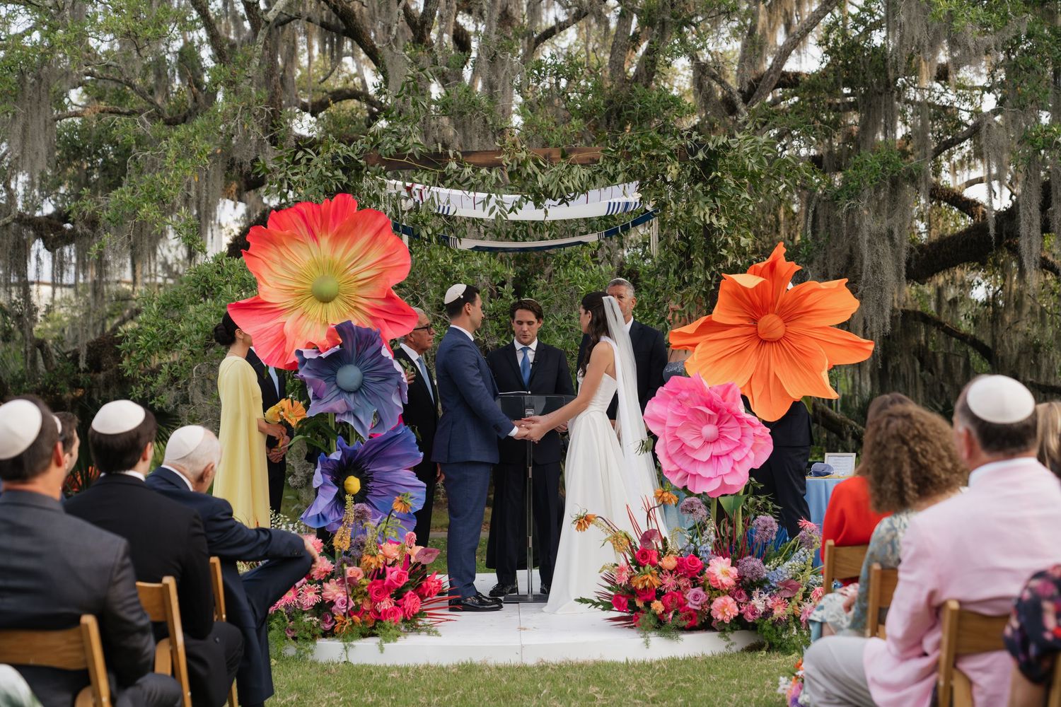 Bride in white gown poses with colorful bridesmaids in rainbow dresses holding bouquets inside a striped venue.
