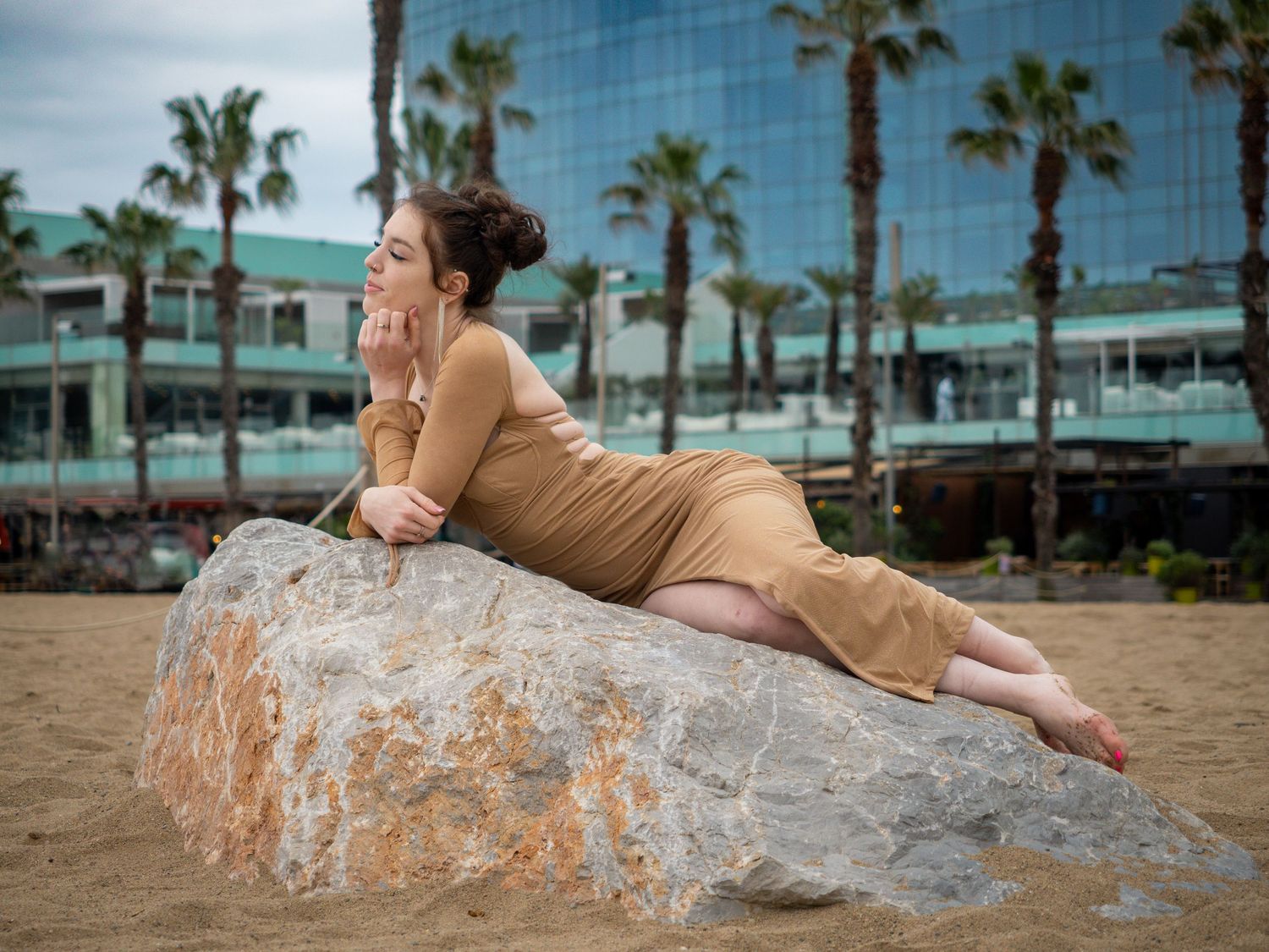 Woman in tan dress poses gracefully on a rock at a sandy beach with palm trees and modern building backdrop.