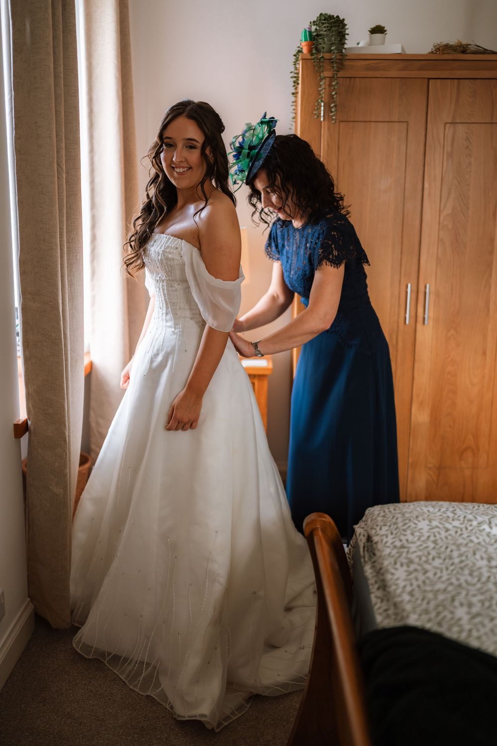 A bride in a white wedding gown gets help with final adjustments in a sunlit room with wooden furniture.