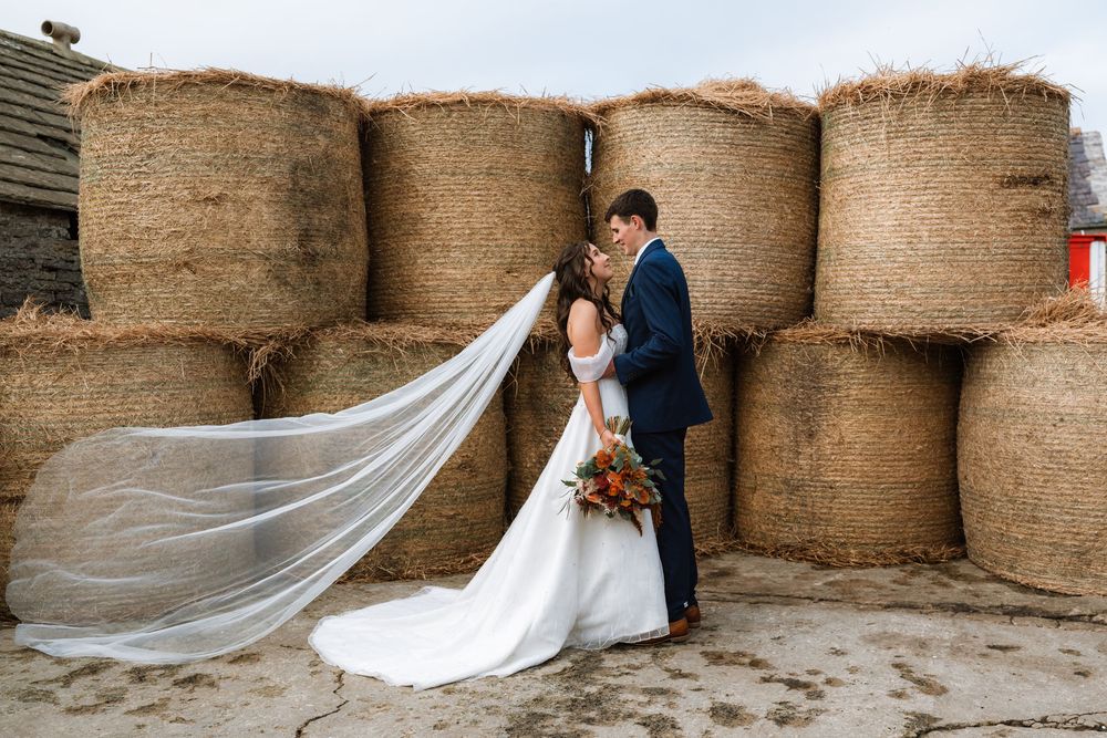 Couple in formal wedding attire embraces against rustic stone wall with flowing white veil creating romantic silhouette.