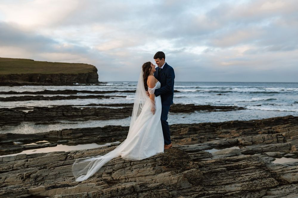 A couple shares an intimate moment on rocky coastal cliffs during sunset.
