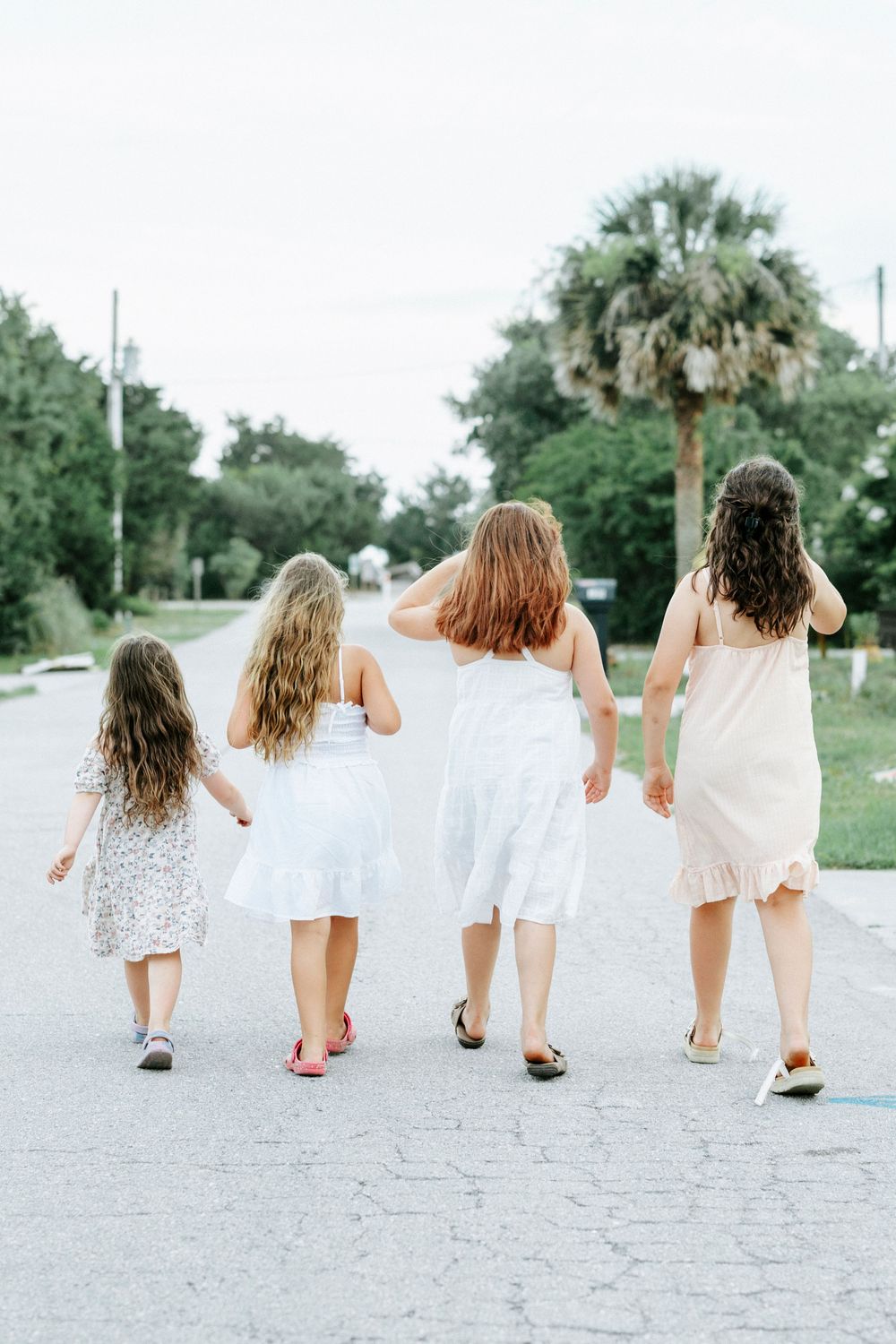 Four girls in white dresses walk together down a tree-lined road, viewed from behind on a cloudy day.