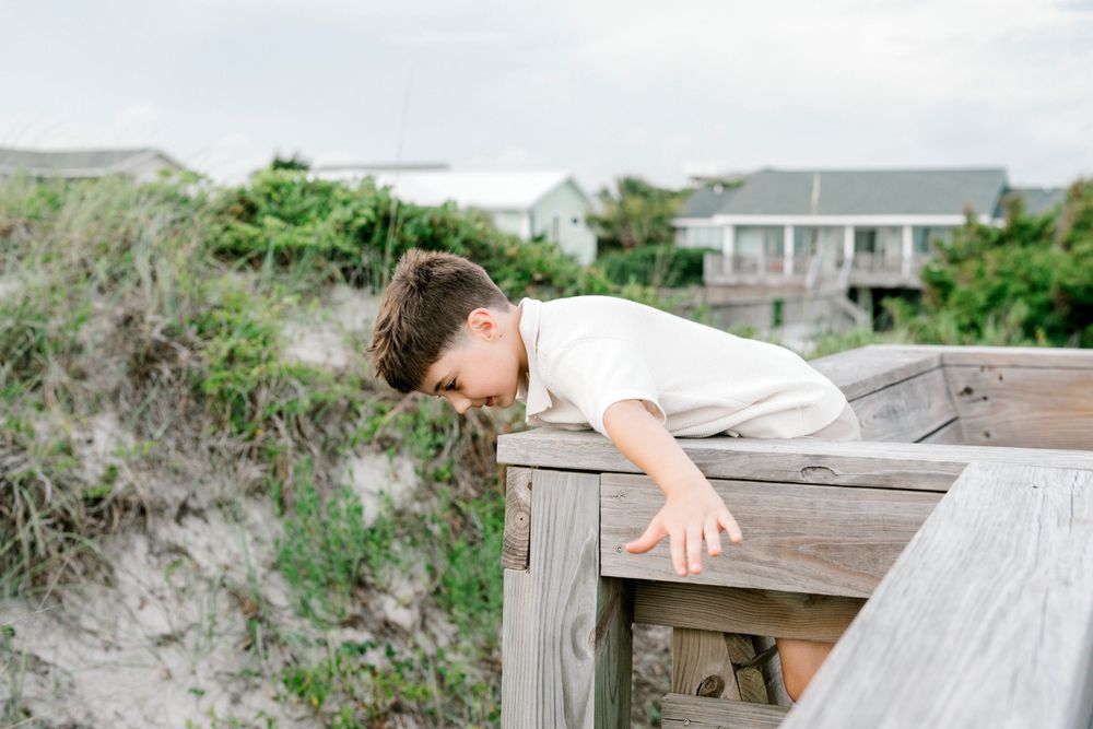 A young boy leans over a wooden boardwalk railing near sandy dunes and beach houses.