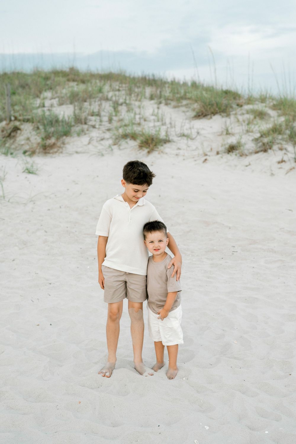 Two young brothers embrace on a sandy beach with dunes and sea grass in the background.