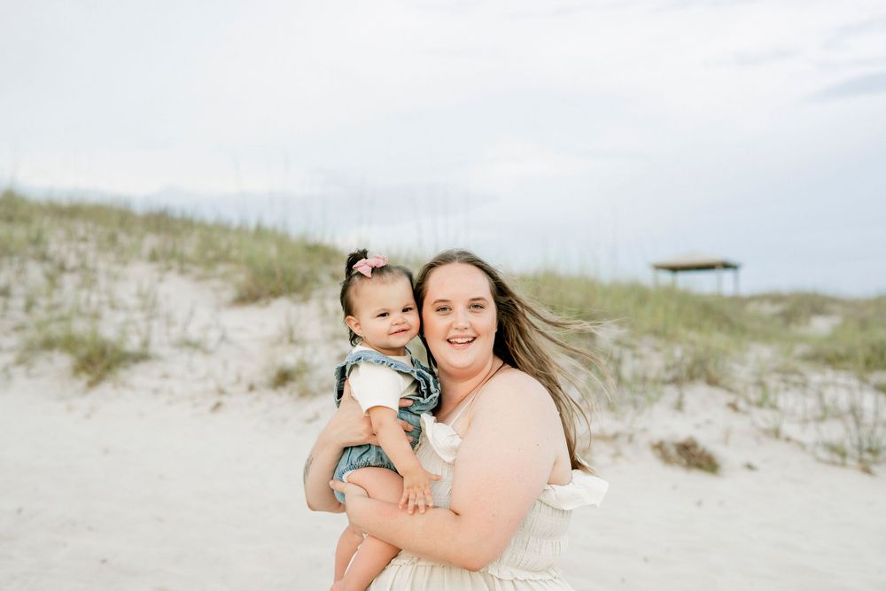 A smiling woman holds a baby girl on a sandy beach with dunes and green grass in the background.
