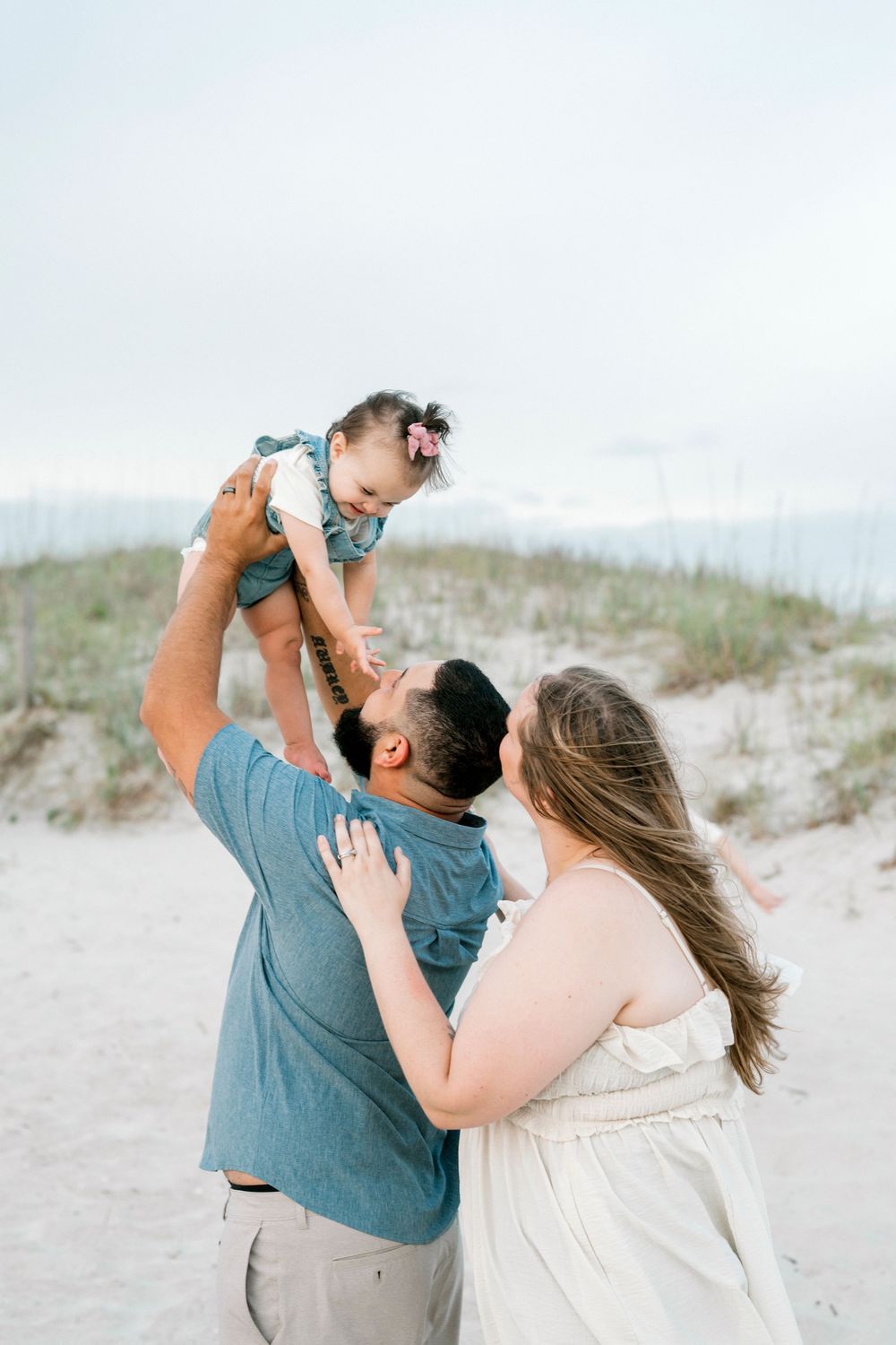 A father lifts his baby girl in the air while mother watches, during a family beach photo session on sandy dunes.