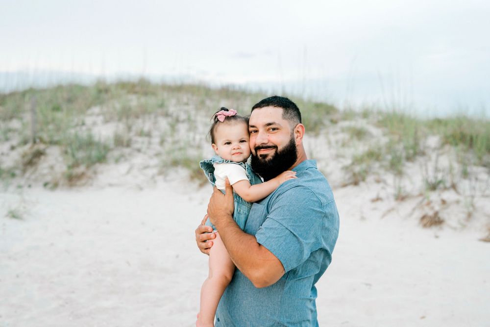 A bearded man holds a smiling toddler girl on a sandy beach with dunes and sea oats in the background.