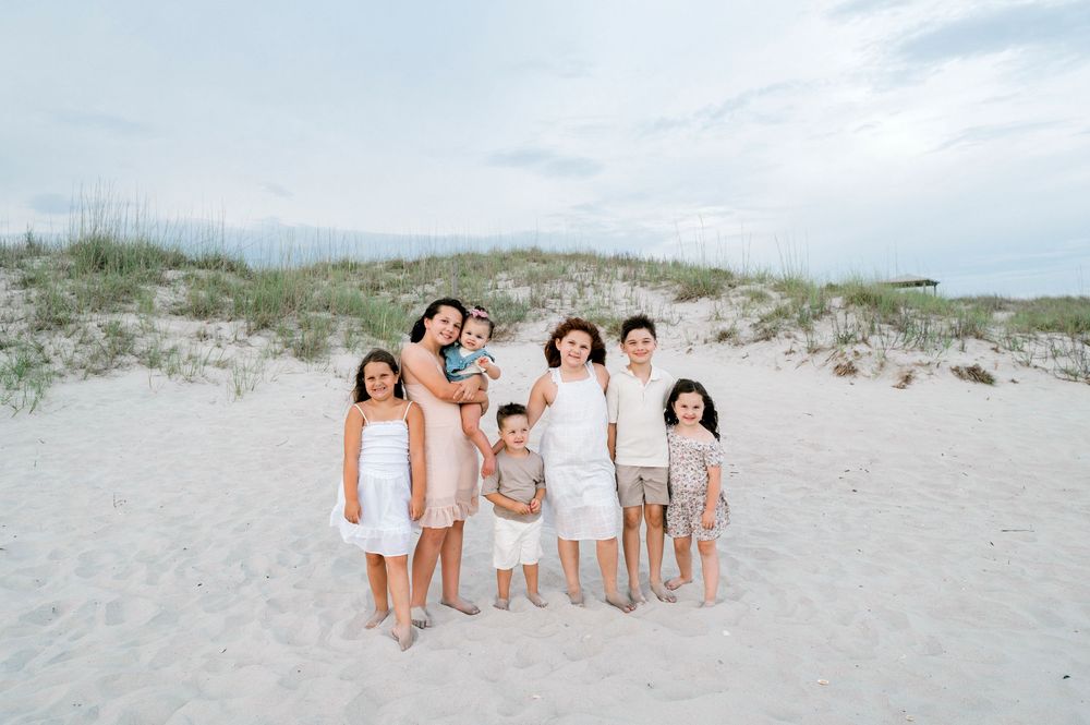 A joyful family of seven poses together on a white sandy beach with sand dunes in the background.